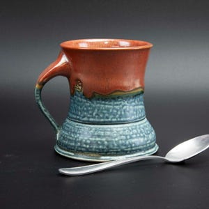 May include: A handcrafted ceramic mug with a textured blue base and a reddish-brown top. The mug has a curved handle and a decorative design near the handle. A silver spoon rests beside the mug on a dark surface.