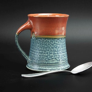 May include: A handmade ceramic mug with a red and blue glaze. The mug has a wide, rounded handle and a flared base.