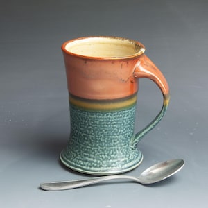 May include: A handmade ceramic mug with a green, yellow, and orange glaze. The mug has a wide base and a handle that curves out from the side. A silver spoon is next to the mug.