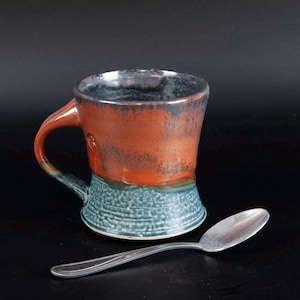 May include: A handmade ceramic mug with a rustic design. The mug features a burnt orange upper half and a textured teal blue lower half. A silver spoon rests beside the mug. The mug has a dark rim and a curved handle.
