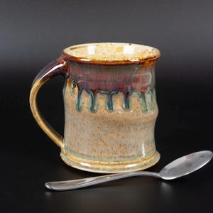May include: A handmade ceramic mug with a brown, blue, and beige glaze. The mug has a unique, slightly uneven shape and a wide, rounded handle.
