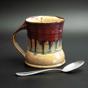 May include: A handmade ceramic mug with a dark red glaze that drips down over a tan base. The mug has a large handle and a silver spoon rests beside it. The mug is likely used for drinking coffee or tea.