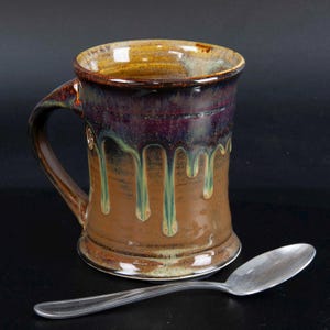 May include: A handmade ceramic mug with a brown, purple, and green glaze. The mug has a handle and a unique drip design. A silver spoon rests beside the mug, suggesting its use for beverages like coffee or tea.