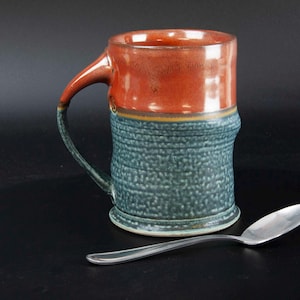 May include: Handmade ceramic mug with a textured teal body and a reddish-brown top. The handle is teal and brown. A silver spoon rests on the black surface next to the mug. This stoneware mug is perfect for coffee or tea.