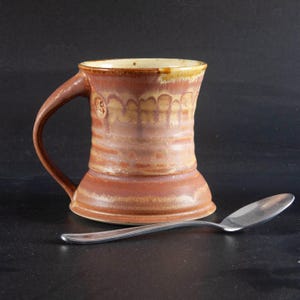 May include: A brown ceramic mug with a wide base and a handle. The mug has a light brown glaze with a yellow rim and a pattern of brown lines.