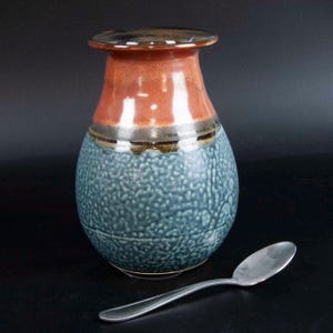 May include: A handcrafted ceramic vase with a textured blue body, a reddish-orange neck, and a dark brown rim. A silver spoon rests beside the vase. The vase has a bulbous shape and a narrow opening. The ceramic vase is a decorative item.