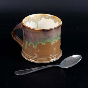 May include: A handmade ceramic mug with a brown base and a colorful glaze of green, purple, and brown. The mug has a handle and a white interior. A silver spoon rests beside the mug.