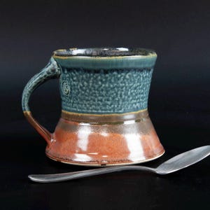May include: A ceramic mug with a flared base, featuring a textured teal upper section and a smooth, glossy reddish-brown lower section. A small circular maker's mark is visible on the side near the handle. A silver metal spoon rests in front.