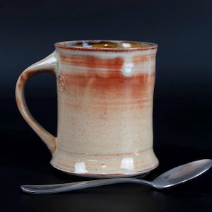 May include: A handmade ceramic mug with a large handle. The mug features a beige base with reddish-brown and white horizontal stripes near the top. A stainless steel spoon rests in front of the mug. The mug is approximately 5 inches tall.