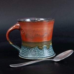 May include: A handmade ceramic mug with a rustic design. The mug features a gradient of colors, including a reddish-orange top, a brown and green mid-section, and a blue base. A silver spoon rests beside the mug.