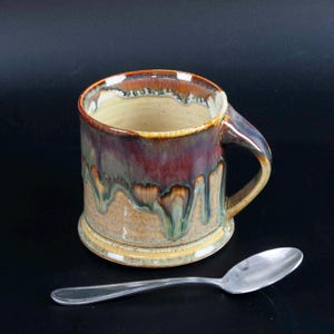 May include: A handmade ceramic mug with a unique glaze in shades of brown, burgundy, and green. The mug has a sturdy handle and a wide base. A silver spoon rests beside the mug, suggesting its use for beverages like coffee or tea.
