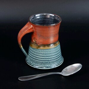 May include: A handmade ceramic mug with a textured, ribbed design. The mug features a burnt orange top, a blue-green base, and a dark interior. A silver spoon rests beside the mug. The mug has a curved handle.