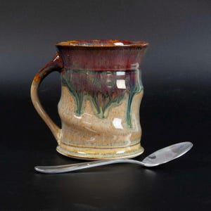 May include: A handmade ceramic mug with a unique, textured design. The mug features a dark burgundy rim, transitioning to green and tan hues. A silver spoon rests beside the mug, set against a black background. This mug is perfect for enjoying coffee or tea.