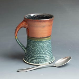 May include: A handmade ceramic mug with a wide, flared base and a large handle. The mug is glazed in shades of red, brown, green, and blue.