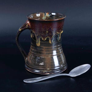 May include: A ceramic mug with a dark brown and black glaze featuring a distinctive drip pattern near the rim, accompanied by a silver-toned metal spoon. The mug has a wide base, a tapered body, and a curved handle on the left side.