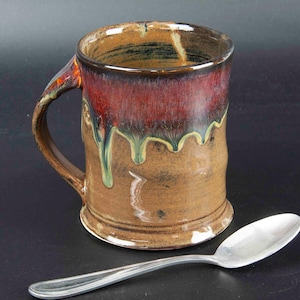May include: A handmade ceramic mug with a brown base and a red-brown top. The mug has a dripped glaze effect with green and yellow accents. A silver spoon rests beside the mug, which has a curved handle.