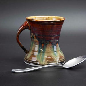 May include: A handmade ceramic mug with a dark brown and green glaze, featuring a unique shape and a sturdy handle. A silver spoon rests beside the mug, suggesting its use for beverages. The mug's design showcases a blend of earthy tones and artistic craftsmanship.