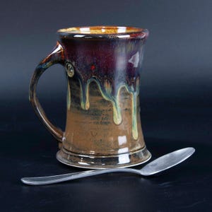 May include: A handcrafted ceramic mug with a unique glaze. The mug features a dark burgundy top that drips down into shades of brown and green. A silver spoon rests beside the mug. The mug has a handle and a decorative button.