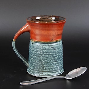 May include: A handmade ceramic mug with a red and blue glaze. The mug has a wide, rounded handle and a slightly flared rim. It is sitting on a black surface next to a silver spoon.