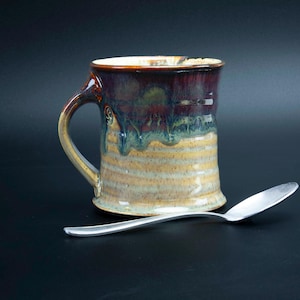 May include: A handmade ceramic mug with a brown, purple, and blue glaze. The mug has a wide, rounded handle and a striped design on the bottom.