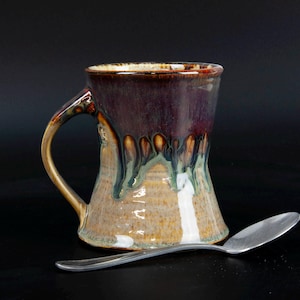May include: A ceramic mug with a flared base and a curved handle, featuring a glossy glaze that transitions from a deep dark brown at the rim to a lighter tan and beige at the bottom with green drip accents. A silver-toned metal spoon lies in front.