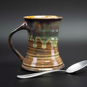 May include: A handcrafted ceramic mug with a unique, textured design. The mug features a brown base, transitioning to green and purple hues towards the top, with a dripped glaze effect. A silver spoon rests beside the mug, suggesting its use for beverages.