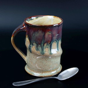 May include: Handmade ceramic mug with a unique glaze. The mug features a brown handle and a body with a blend of colors, including tan, burgundy, and green. A silver spoon rests beside the mug, suggesting its use for beverages like coffee or tea.