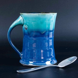 May include: A handcrafted ceramic mug with a blue and turquoise ombre glaze. The mug features a curved handle and a small logo near the handle. A silver spoon rests beside the mug.