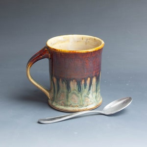 May include: A brown and green ceramic mug with a handle. The mug has a drip glaze design and is sitting on a gray surface with a silver spoon.