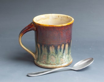 Może przedstawiać: Brązowy i zielony kubek ceramiczny z uchwytem. Kubek ma wzór glazury w kroplach i stoi na szarym tle z srebrną łyżeczką.