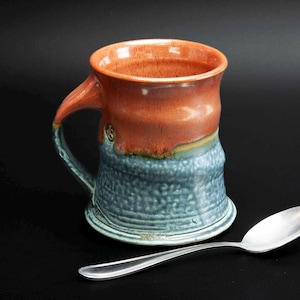 May include: A handmade ceramic mug with a blue and red glaze. The mug has a unique, slightly uneven shape and a wide, rounded handle.