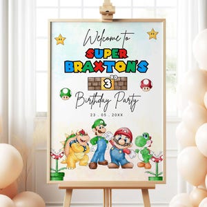 May include: A wooden easel holds a birthday party sign with the text "Welcome to Super Braxton's 3rd Birthday Party." The sign features cartoon characters and star and mushroom graphics. Beige balloons are clustered around the easel.