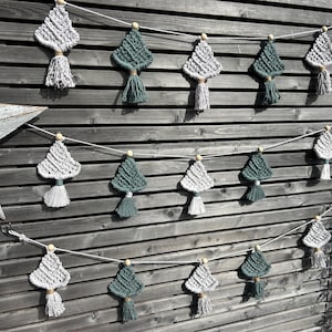May include: A macramé garland featuring diamond-shaped ornaments in shades of grey and green, each with a tassel. The garland is strung on a white cord and accented with wooden beads. The background is a wooden fence.