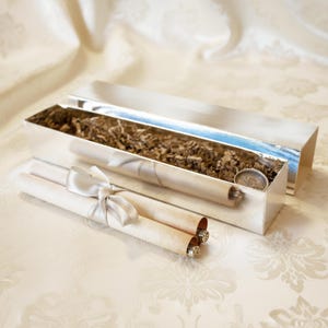 May include: A silver rectangular box with a lid, filled with brown packing material. Two rolled scrolls tied with a white satin ribbon are in front of the box. A small silver coin is inside the box.
