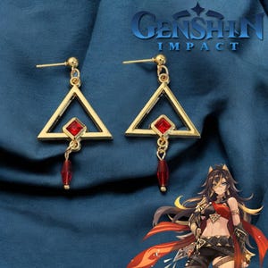 Può includere: Orecchini a forma di triangolo dorati con accenti di pietre preziose rosse. Gli orecchini sono pendenti e hanno una piccola pietra preziosa rossa appesa alla parte inferiore. Gli orecchini sono ispirati al videogioco Genshin Impact.