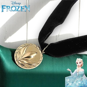 May include: A gold-colored pendant necklace with a black velvet ribbon. The pendant has a leaf design and is round. The Disney Frozen logo is in the upper left corner.