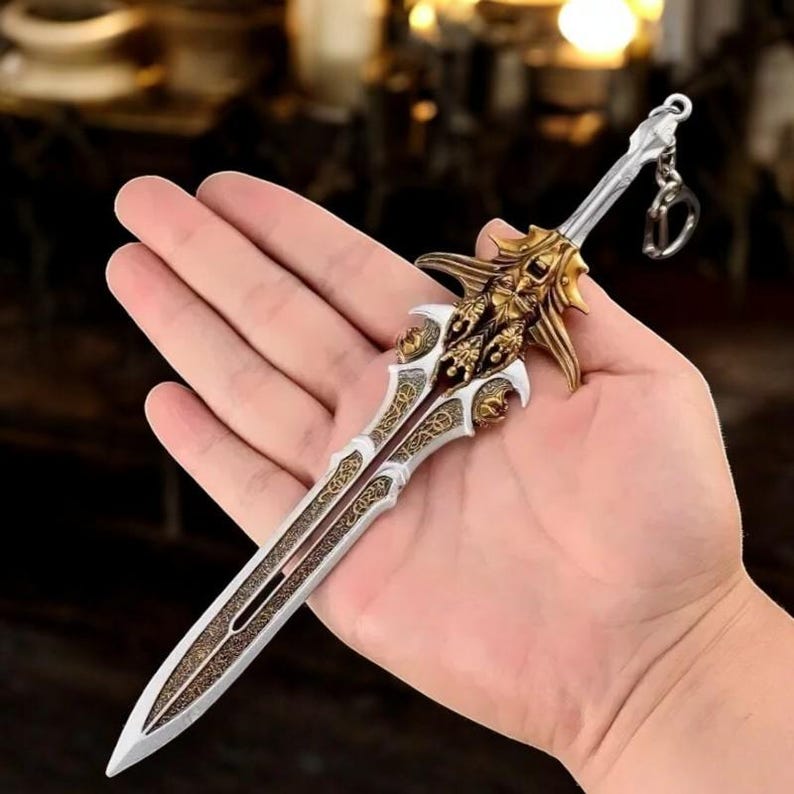May include: A miniature decorative sword keychain. The sword features a silver blade with gold filigree and a gold hilt with intricate details. A small silver keyring is attached to the hilt.