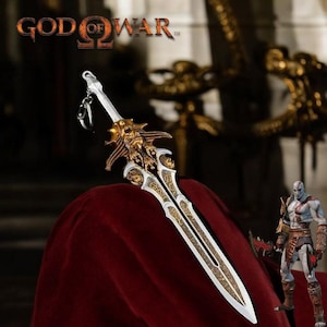 May include: A detailed sword keychain with a gold and silver design, featuring intricate patterns and a skull-like emblem. The keychain is displayed on a red surface, with the "God of War" logo visible at the top. A Kratos action figure is also in the image.