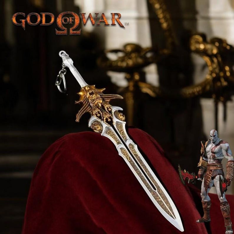 May include: A detailed sword keychain with a gold and silver design, featuring intricate patterns and a skull-like emblem. The keychain is displayed on a red surface, with the "God of War" logo visible at the top. A Kratos action figure is also in the image.