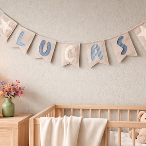 May include: A banner spelling "LUCAS" hangs above a wooden cot. The letters are in blue and cream, with star-shaped accents on each end. A teddy bear peeks out from the cot. A lamp and vase of flowers sit on a wooden chest of drawers.