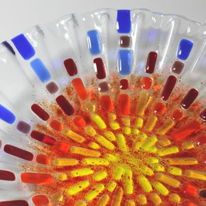 May include: A clear glass bowl with a decorative design of red, orange, yellow, and blue glass pieces. The pieces are arranged in a radial pattern, creating a sunburst effect.