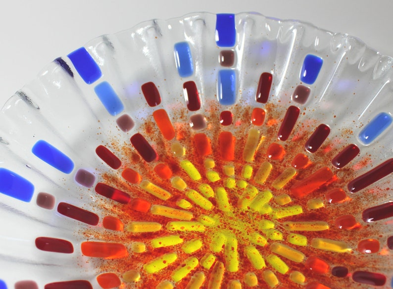 May include: A clear glass bowl with a decorative design of red, orange, yellow, and blue glass pieces. The pieces are arranged in a radial pattern, creating a sunburst effect.