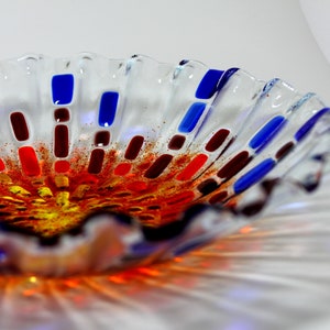 May include: A clear glass bowl with a scalloped edge. The bowl is decorated with red, orange, yellow, brown, and blue glass pieces.