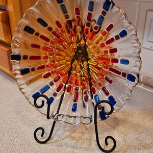May include: A clear glass plate with a scalloped edge and a colorful mosaic design. The mosaic features red, orange, yellow, blue, and brown glass pieces. The plate is on a black metal stand.