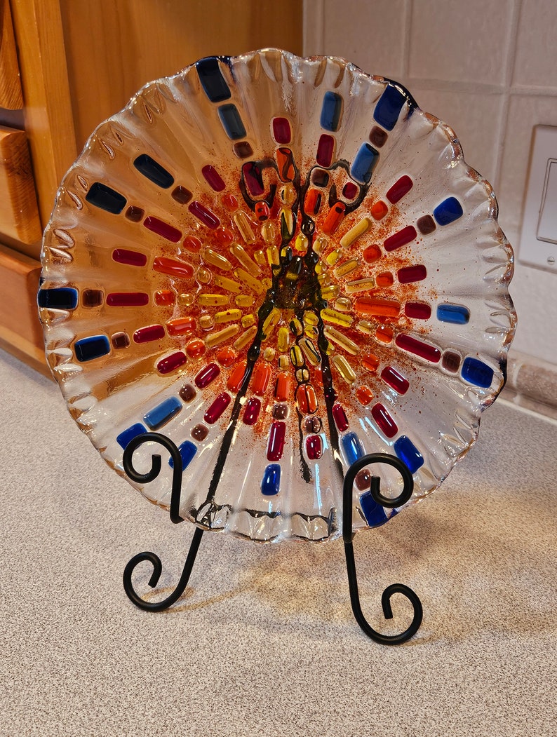 May include: A clear glass plate with a scalloped edge and a colorful mosaic design. The mosaic features red, orange, yellow, blue, and brown glass pieces. The plate is on a black metal stand.