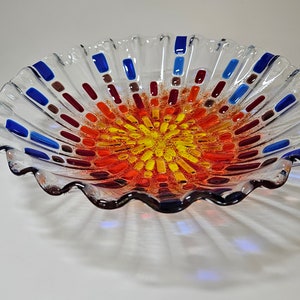 May include: A clear glass bowl with a wavy edge and a colorful mosaic design in the center. The mosaic features shades of red, orange, yellow, and blue.