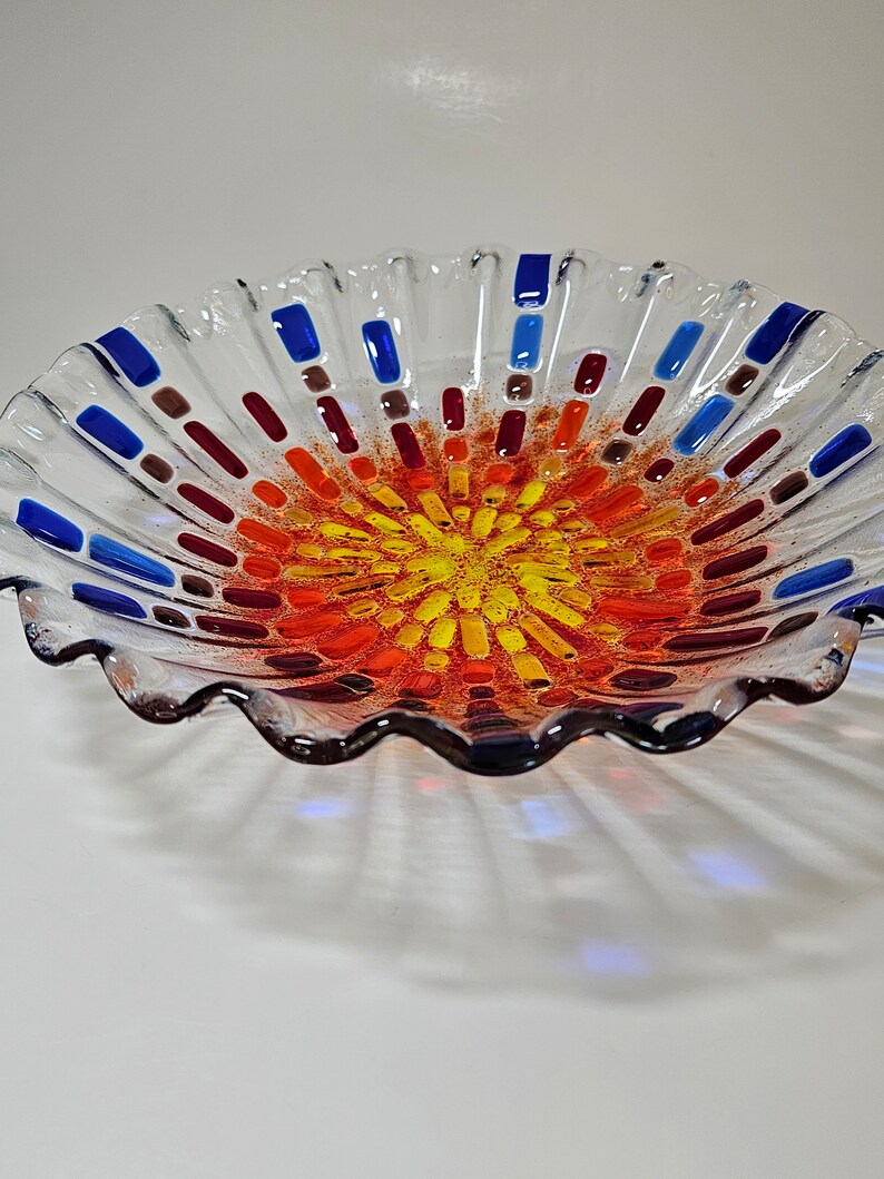 May include: A clear glass bowl with a wavy edge and a colorful mosaic design in the center. The mosaic features shades of red, orange, yellow, and blue.