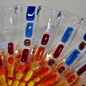 May include: A clear glass bowl with a textured surface. The bowl is decorated with small pieces of colored glass in shades of red, orange, yellow, and blue. The glass pieces are arranged in a circular pattern, creating a sunburst effect.