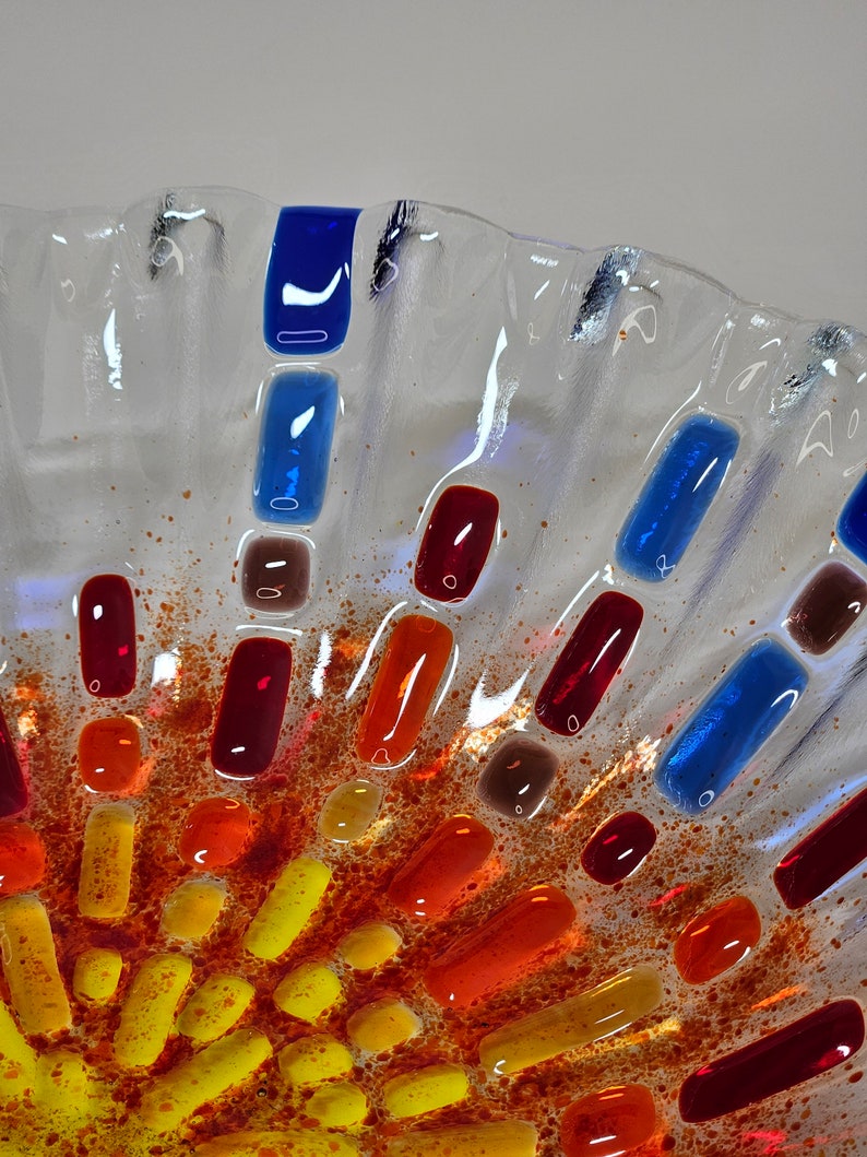 May include: A clear glass bowl with a textured surface. The bowl is decorated with small pieces of colored glass in shades of red, orange, yellow, and blue. The glass pieces are arranged in a circular pattern, creating a sunburst effect.
