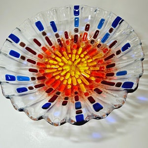 May include: A clear glass bowl with a scalloped edge and a colorful mosaic design in the center. The mosaic features shades of red, orange, yellow, blue, and brown.